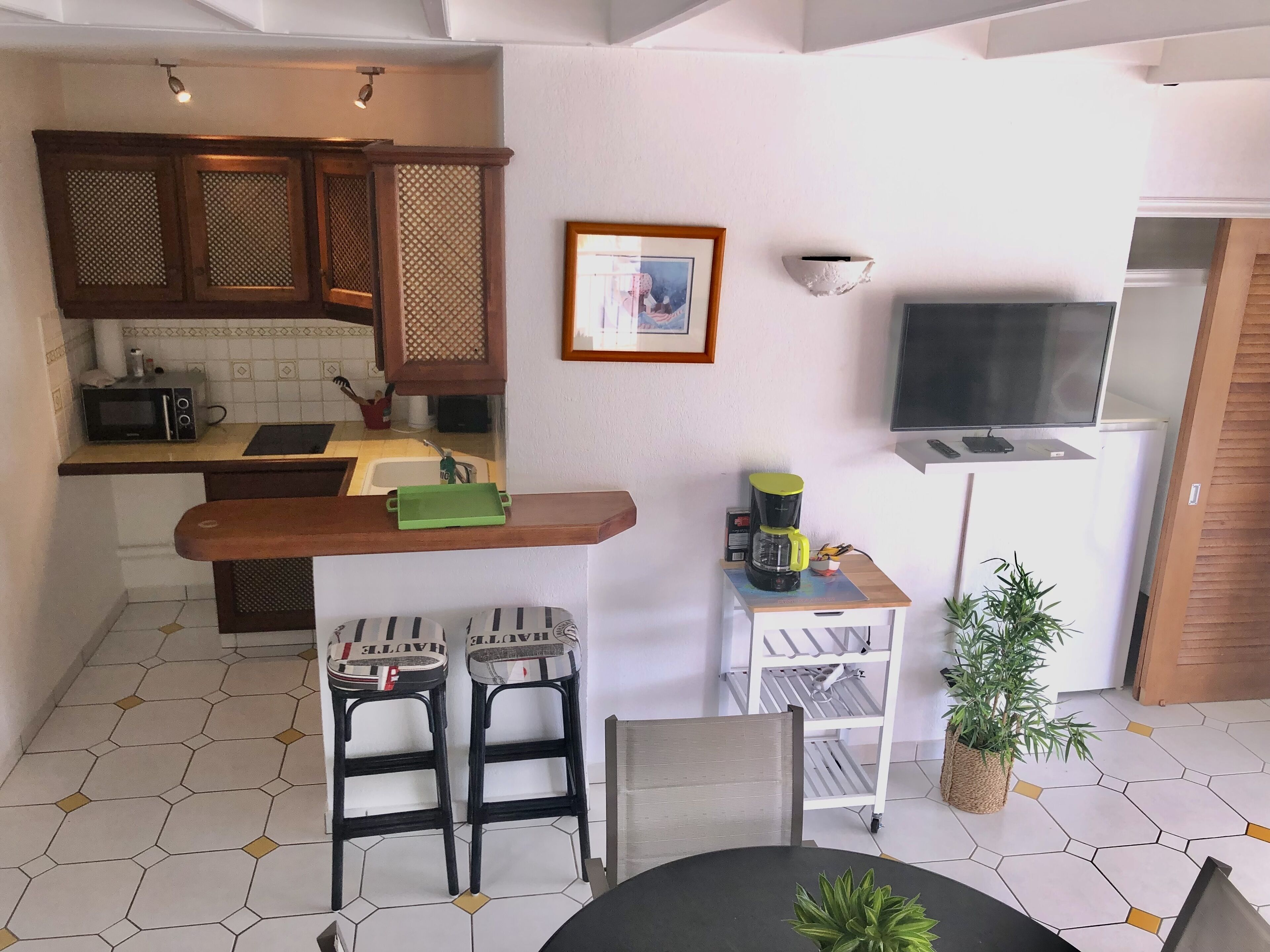 A compact kitchen with a microwave, toaster, and sink. There are two bar stools, a small table, and a potted plant. A framed picture hangs on the wall, and a TV is mounted above a white cabinet.