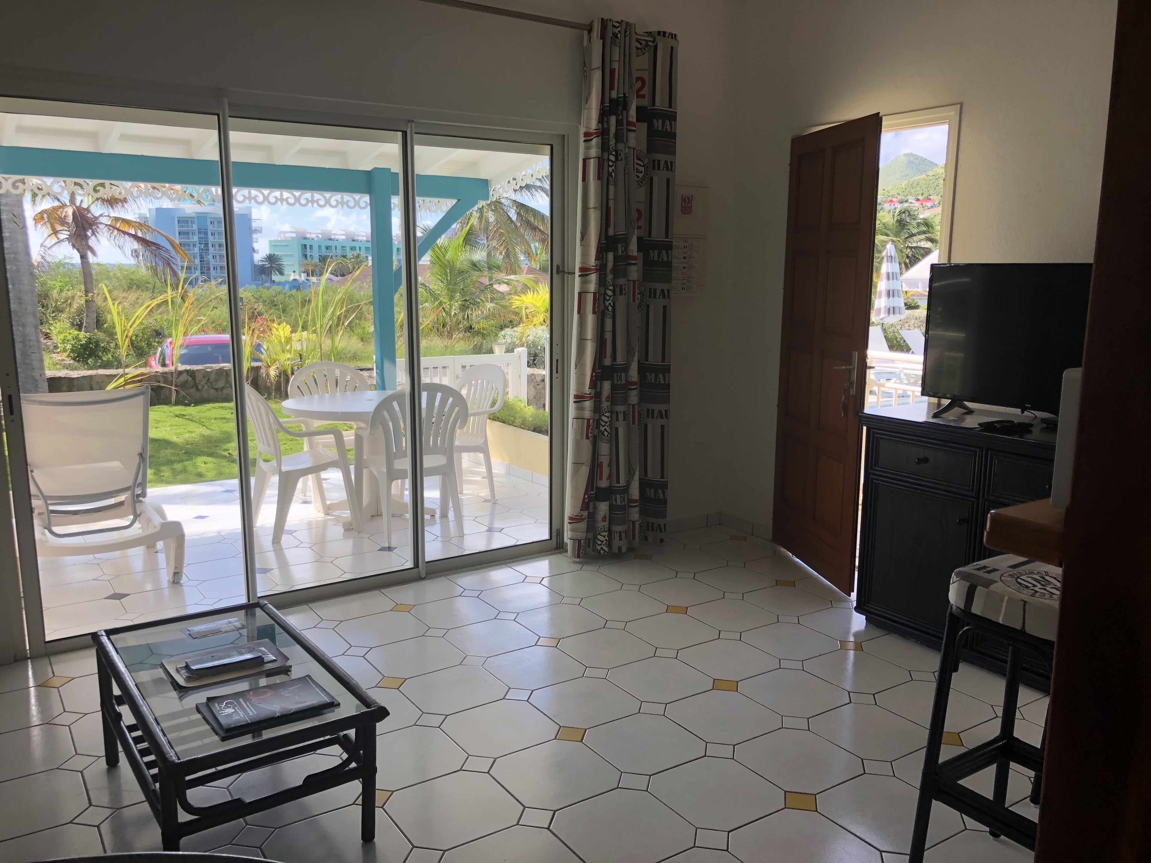 A room with a glass door leading to an outdoor area with chairs and a table. There is a TV on a cabinet and a window with a view of greenery and buildings.
