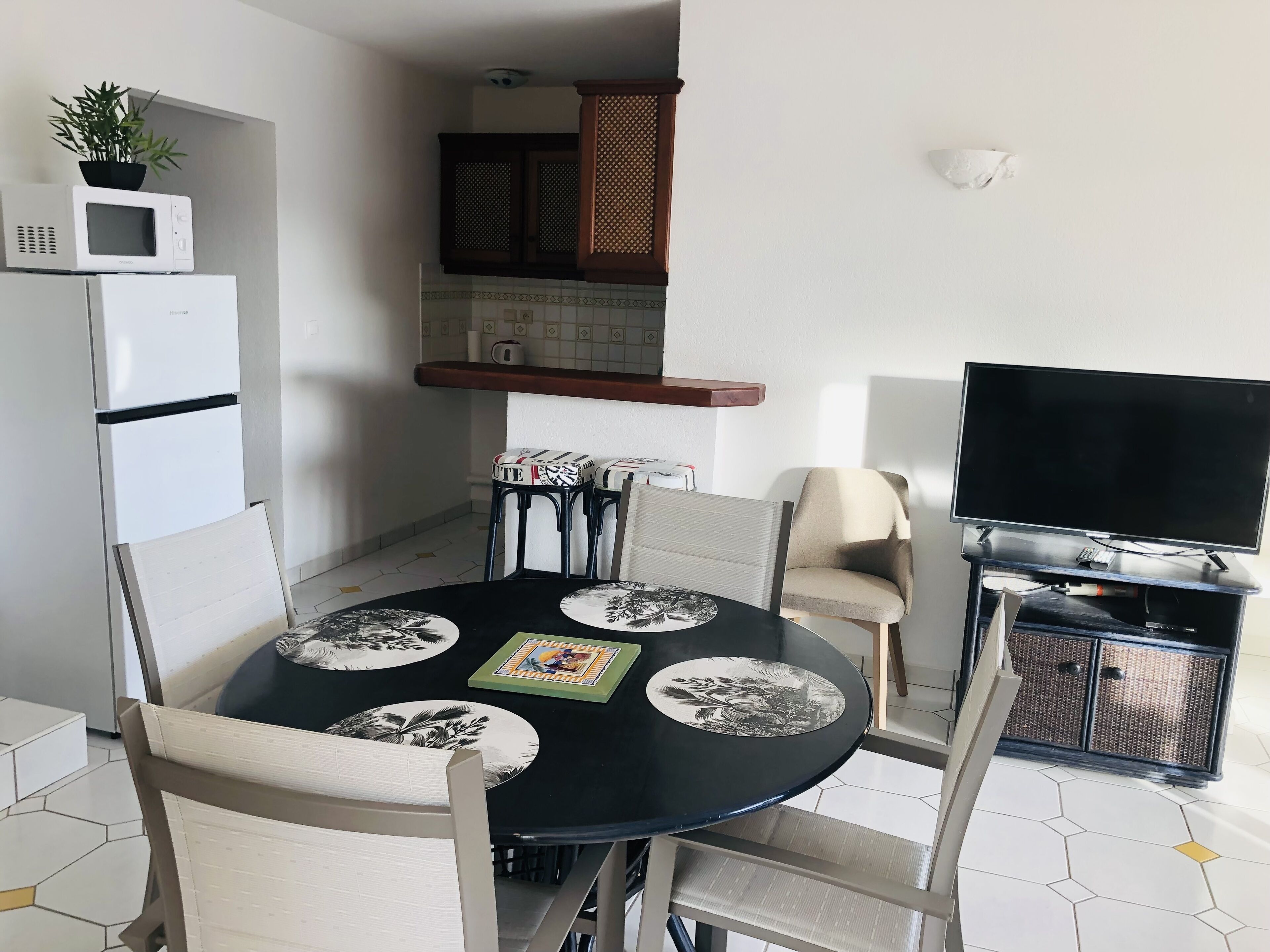 Un espacio habitable compacto con una mesa de comedor rodeada de sillas, un microondas, un refrigerador, un televisor y una pequeña cocina con armarios y un estante.