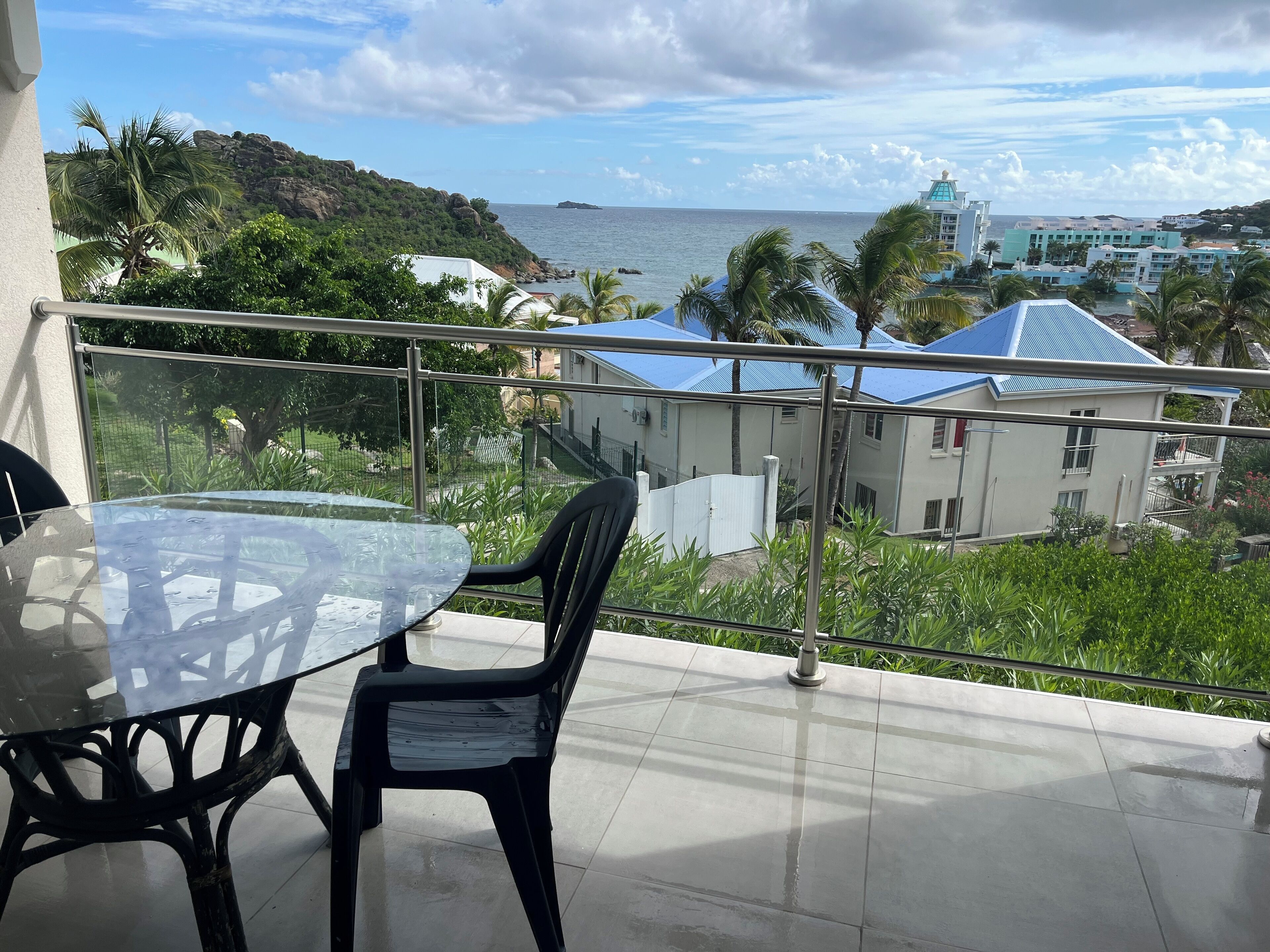 Un balcon doté d’une table et de chaises en verre, offrant une vue sur une zone côtière avec des bâtiments et de la végétation.