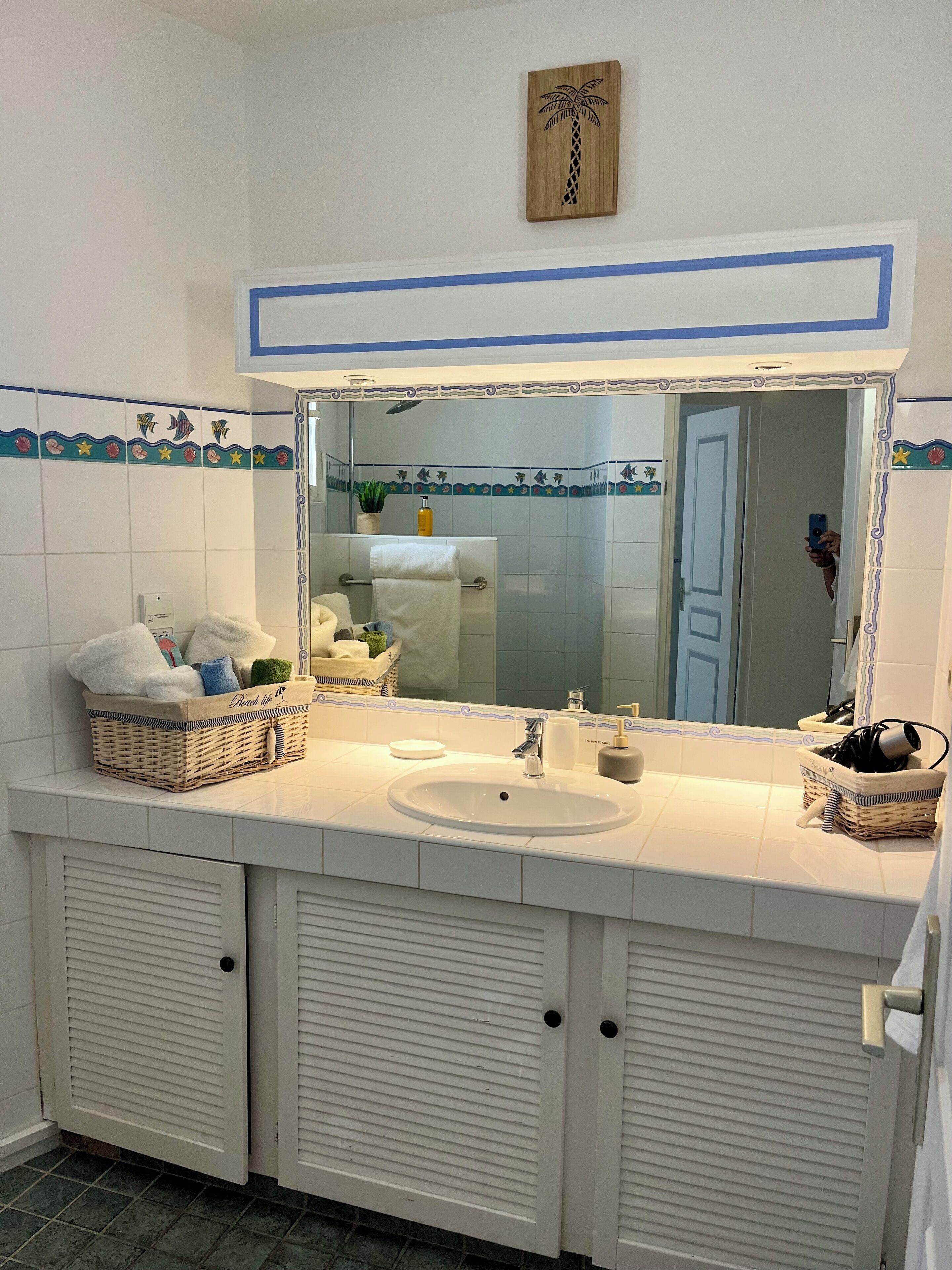 Ein Badezimmer mit einem weißen Waschbecken, einem Spiegel und zwei Schränken. Ein Handtuchhalter mit einem weißen Handtuch und ein Korb mit Pflegeprodukten.