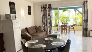 A dining area with a round table, chairs, and a refrigerator. There is a sofa and a view of a balcony with curtains. - Colombus Résidence (Oyster Pond)