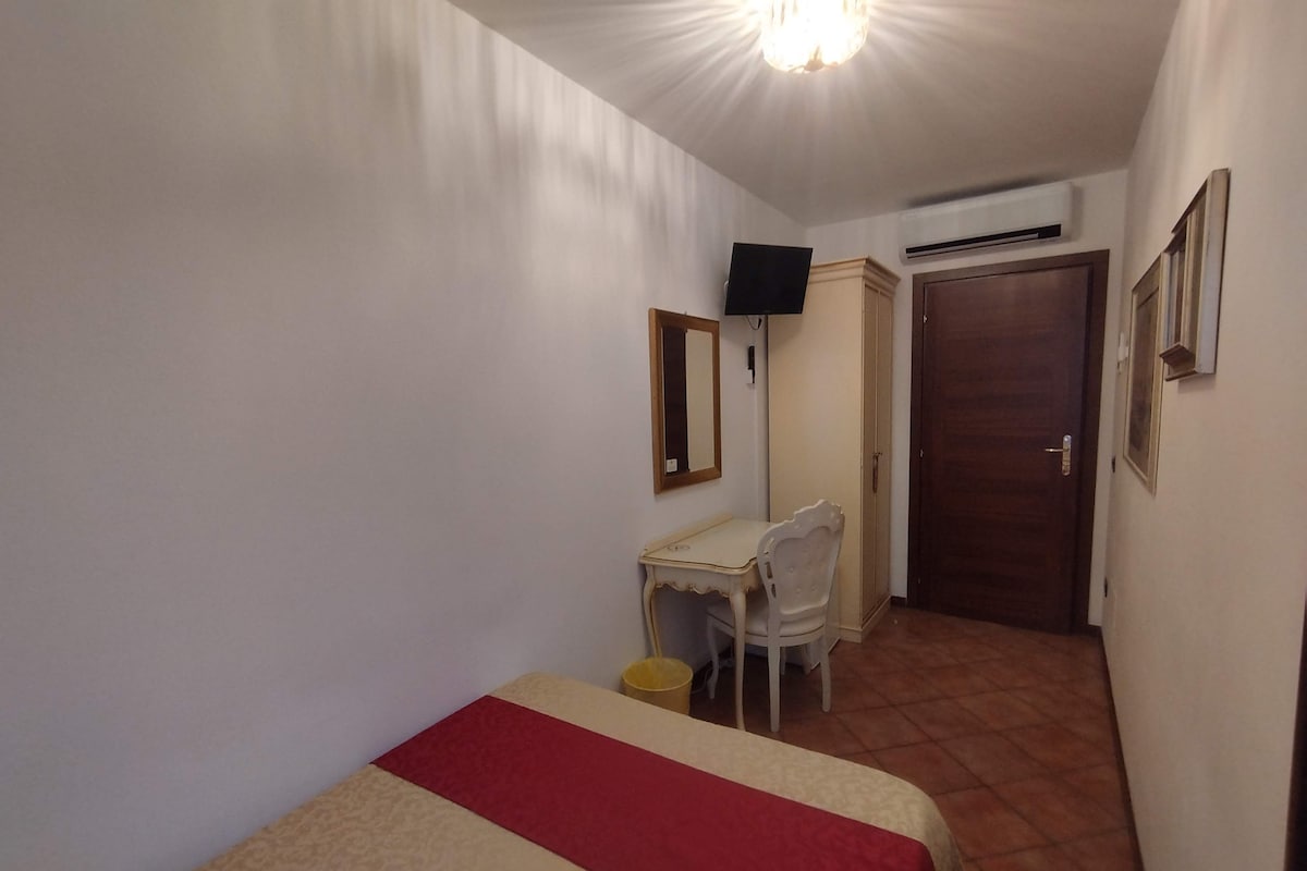 A small room with a bed, a desk, a chair, a television, and an air conditioning unit.