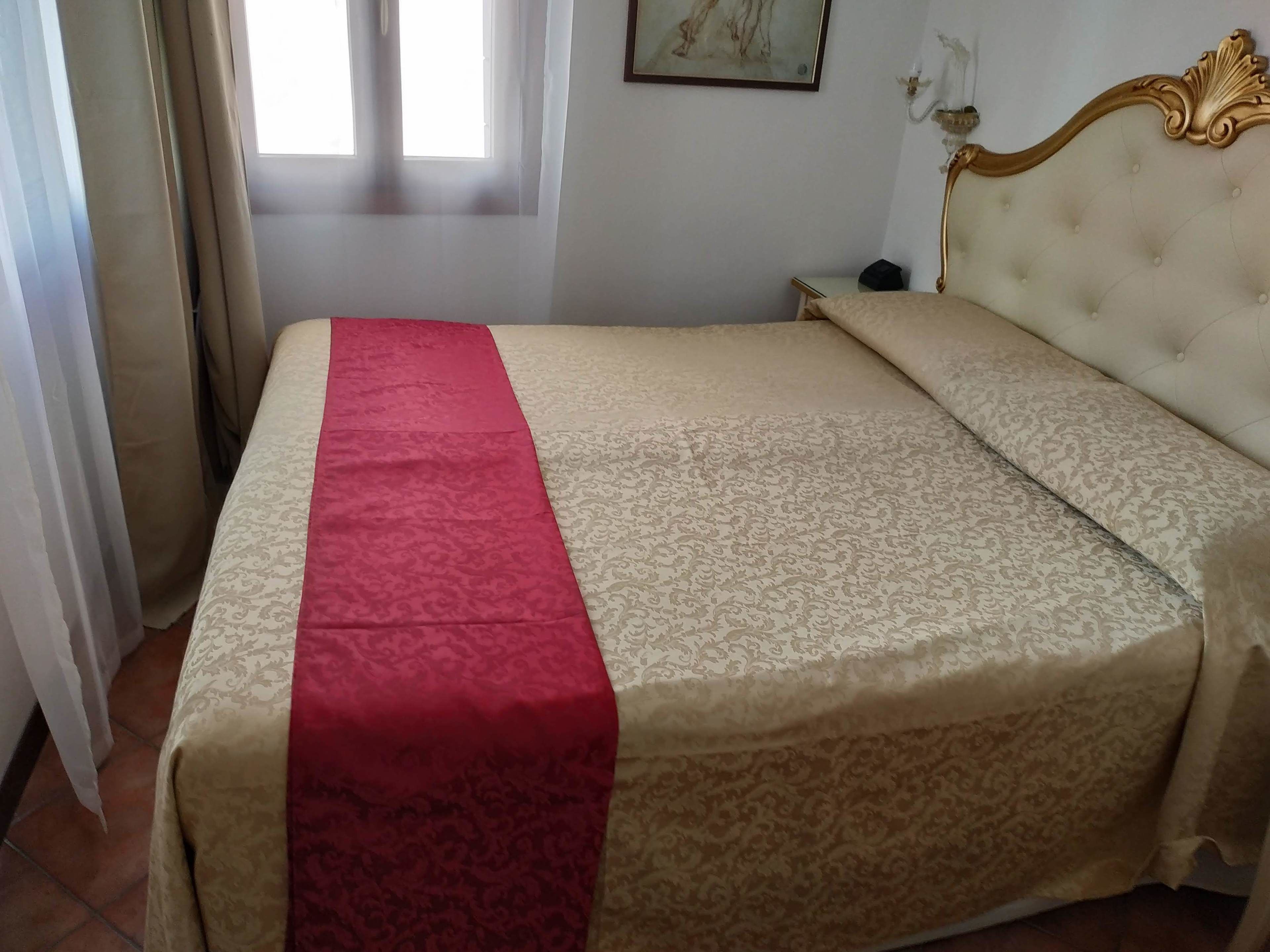 Een net opgemaakt bed met een rood-beige dekbed, een gouden hoofdeinde en een nachtkastje met een lamp.