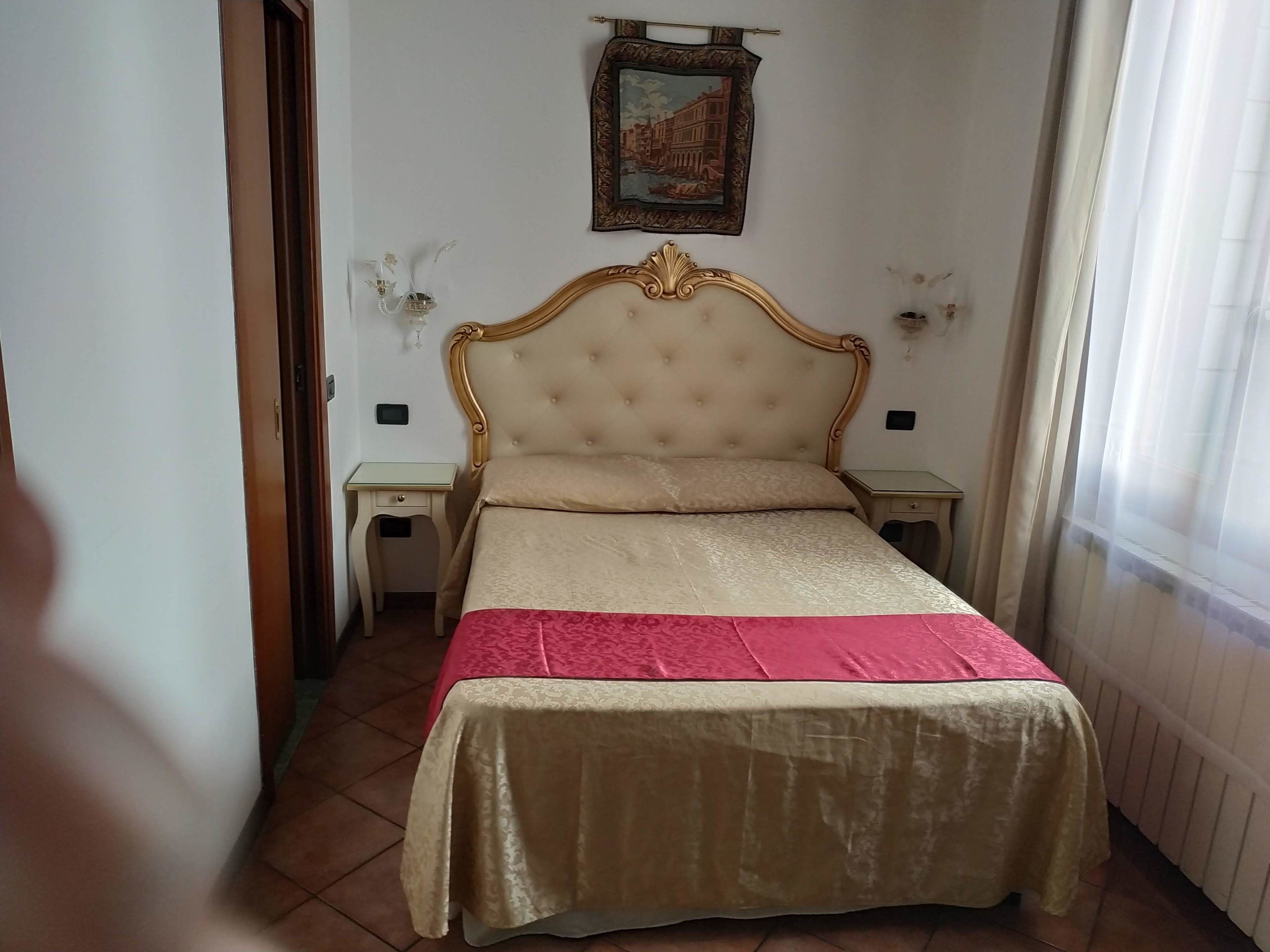 A bedroom with a tufted headboard, a bed with a red and gold bedspread, two bedside tables with lamps, and a framed picture on the wall.