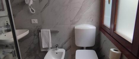 Een badkamer met een bidet, toilet en wastafel. Boven het toilet bevindt zich een handdoekhouder, met een handdoek erop. Een haardroger is aan de muur bevestigd.