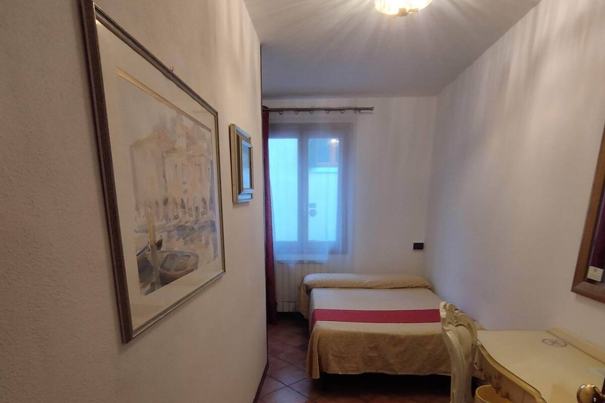 A small, compact room with a bed, a desk, and a chair. There is a window with curtains, a door, and a wall-mounted light fixture.