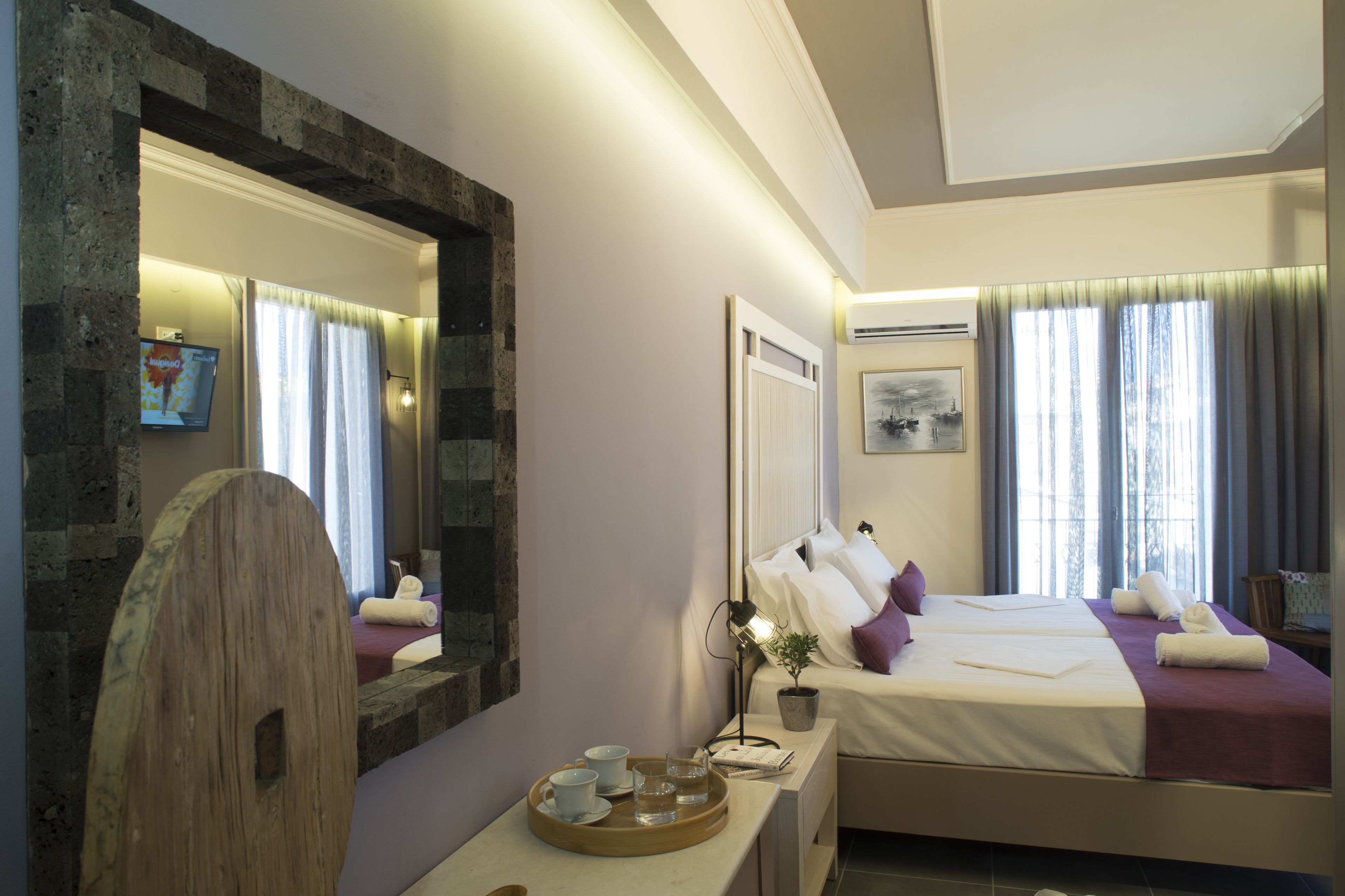 A hotel room with a large mirror, two beds, a bedside table with a tray of cups and glasses, a potted plant, and a painting on the wall.