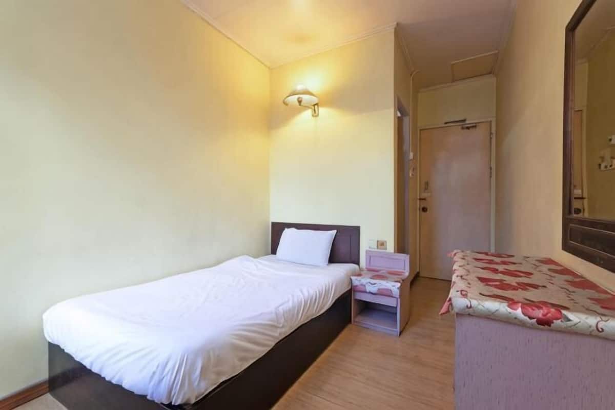 A single bed with a white mattress, a bedside table with a lamp, and a door leading to a bathroom.