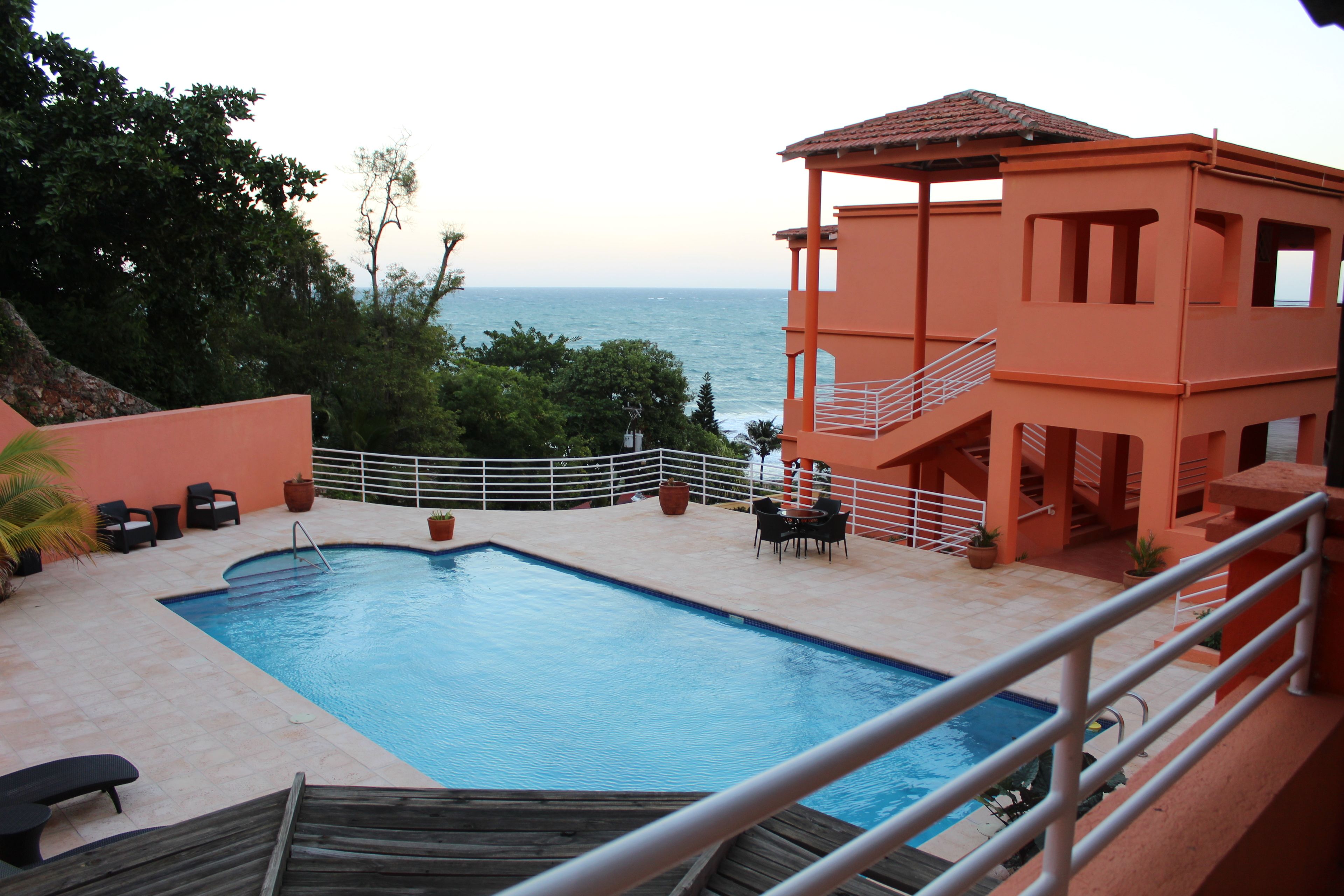 A swimming pool with a view of the sea, surrounded by outdoor furniture and a building with a balcony.