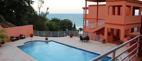 A swimming pool with a view of the sea, surrounded by outdoor furniture and a building with a balcony.