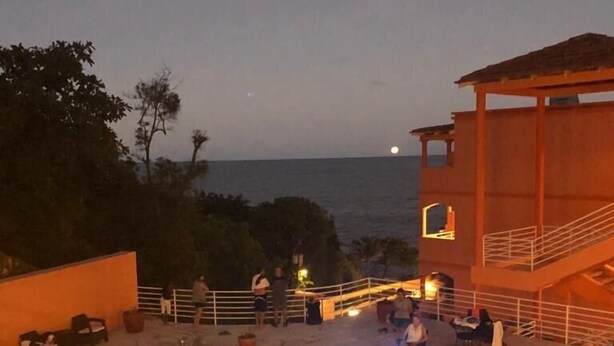 A swimming pool with people in it, surrounded by a building with a balcony and a view of the sea. - Rival Hotel (Cap-Haitien)