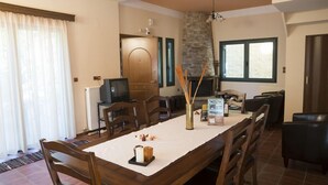 A dining area with a long table, chairs, a television, and a stone wall. - Elia Therissos Maisonettes (Chania)
