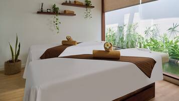 Habitación de hotel con dos camas, un ventanal con vistas a zonas verdes, suelo de madera y un estante en la pared con plantas y toallas.