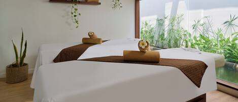 Habitación de hotel con dos camas, un ventanal con vistas a zonas verdes, suelo de madera y un estante en la pared con plantas y toallas.