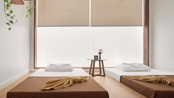 A minimalist room with two beds, a small table with a clock, and a window with blinds.