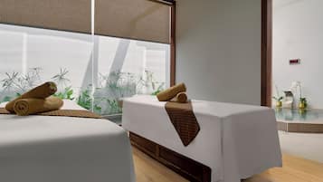 A modern spa room with two massage tables, a large window with plants, and a digital clock.