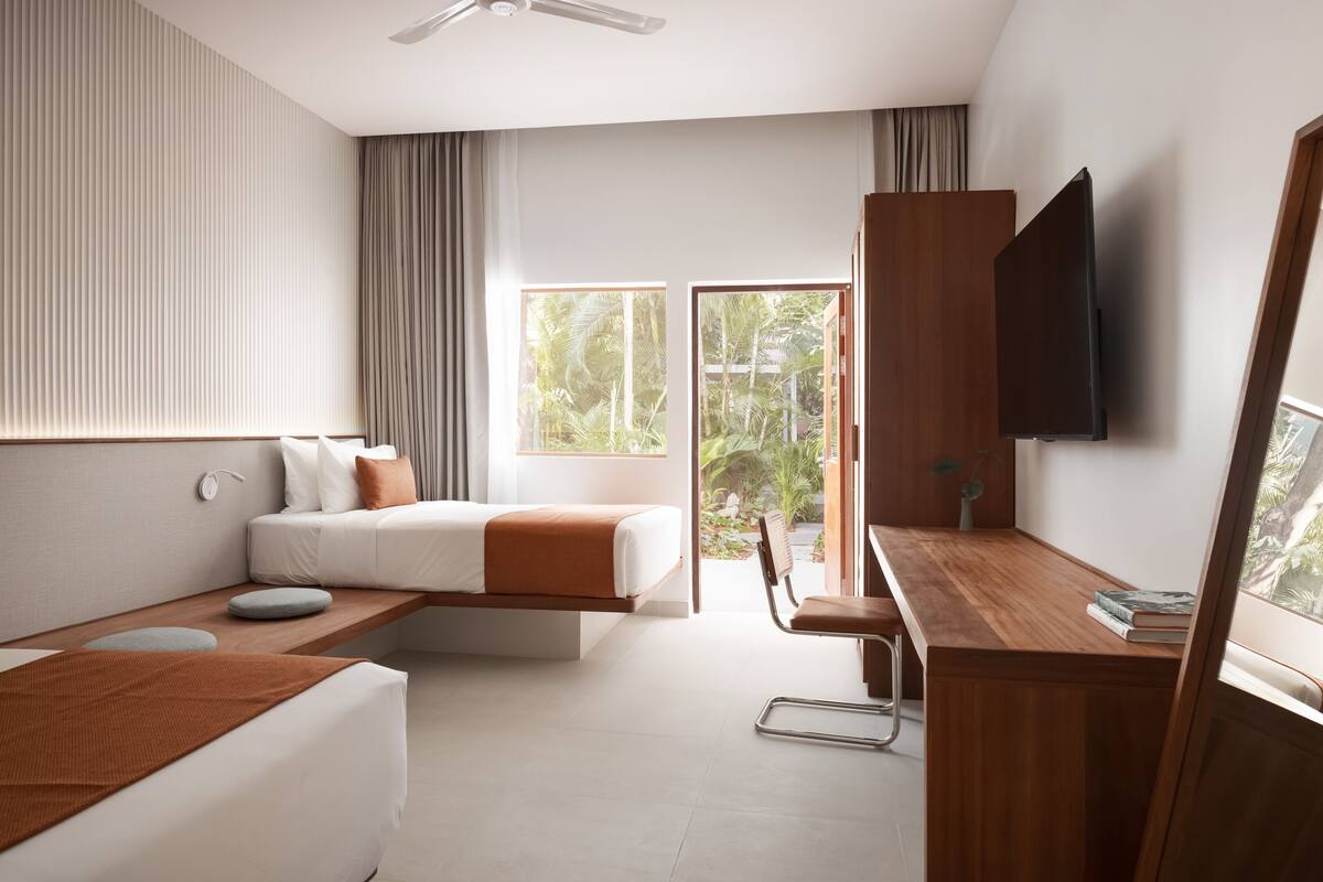 Ein modernes Hotelzimmer mit Bett, Schreibtisch, Stuhl, Fernseher und großen Fenstern mit Blick auf Grünflächen.
