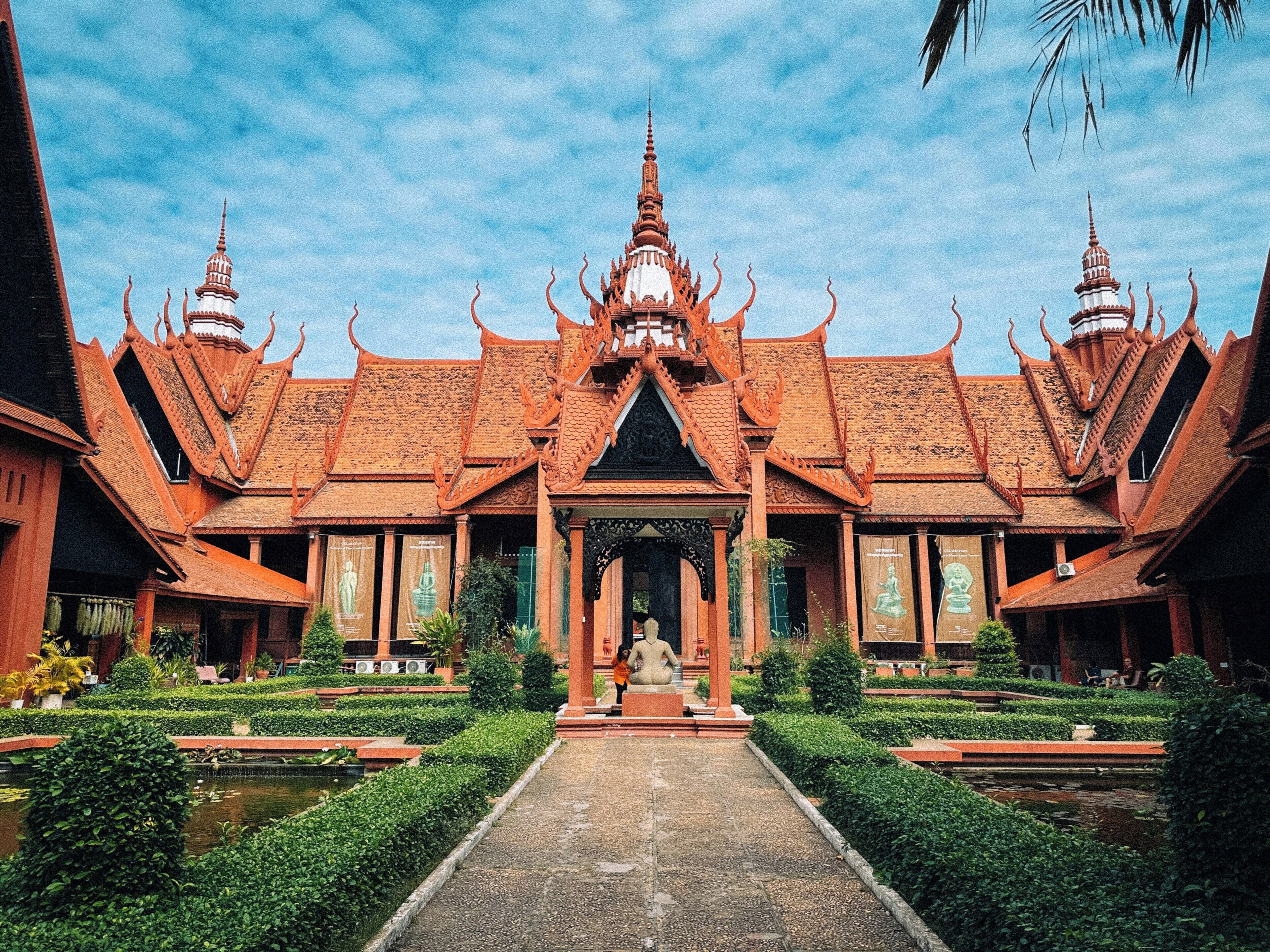 A traditional Thai temple with a central walkway leading to a statue, surrounded by symmetrical architecture and well-maintained gardens.