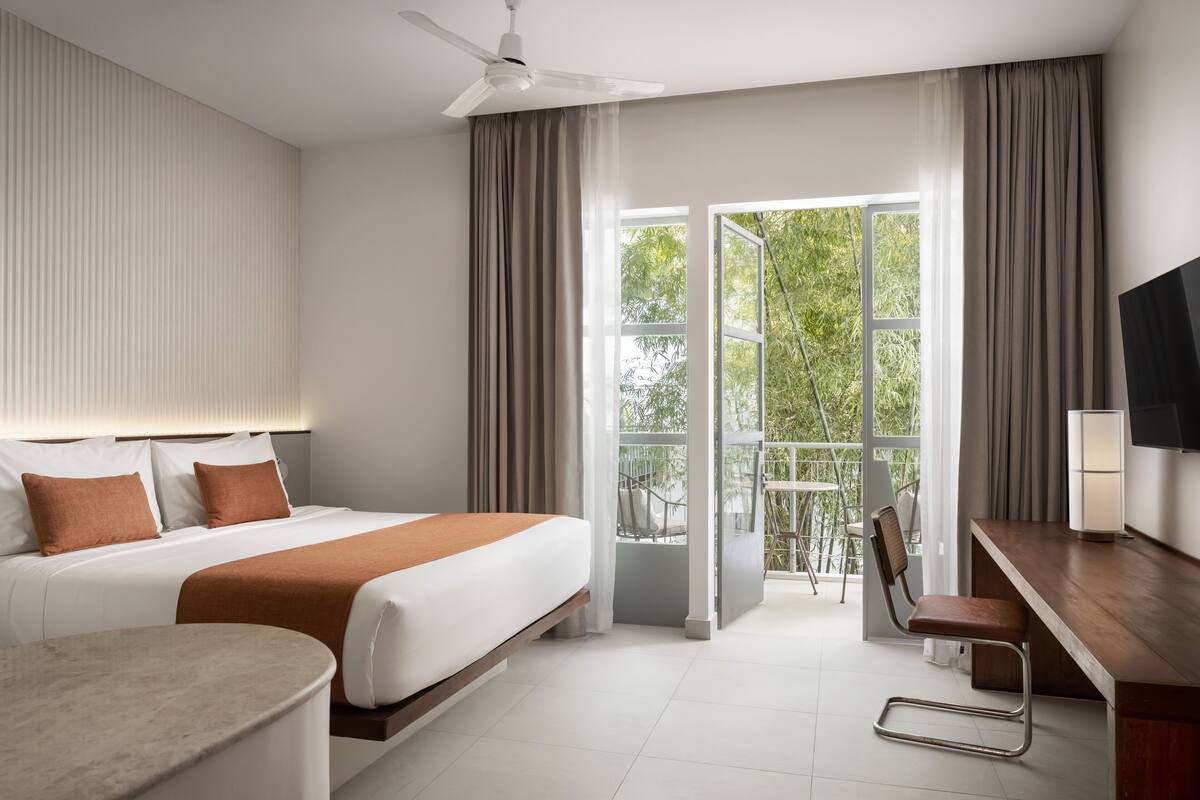Ein modernes Hotelzimmer mit Bett, Schreibtisch, Stuhl und einem Balkon mit Blick auf Grünflächen.