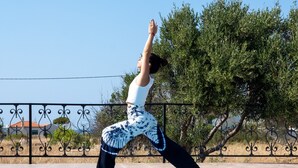 A person performing a yoga pose on a blue mat by a swimming pool with a clear sky and trees in the background. - Elia Stavros Villas (Chania)
