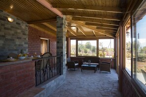 Un patio couvert doté d’un sol en pierre, de poutres en bois au plafond, de murs en briques et de grandes fenêtres offrant une vue sur l’extérieur.