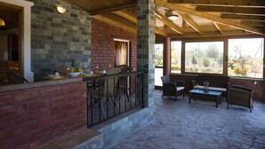 A covered patio area with stone flooring, brick walls, and wooden beams. There are outdoor seating arrangements with chairs and a table, and a window providing a view of the outdoors. - Elia Stavros Villas (Chania)