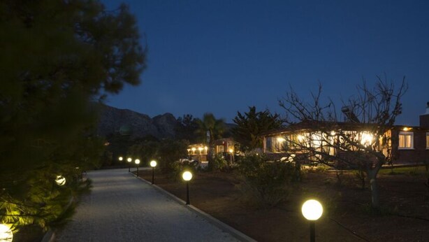A pathway leading to a building with multiple lit windows, surrounded by trees and a mountain in the background. - Elia Stavros Villas (Chania)