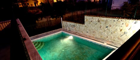 Une piscine avec un mur en pierre et un bord carrelé, entourée d’une clôture et bordée de bâtiments en arrière-plan.