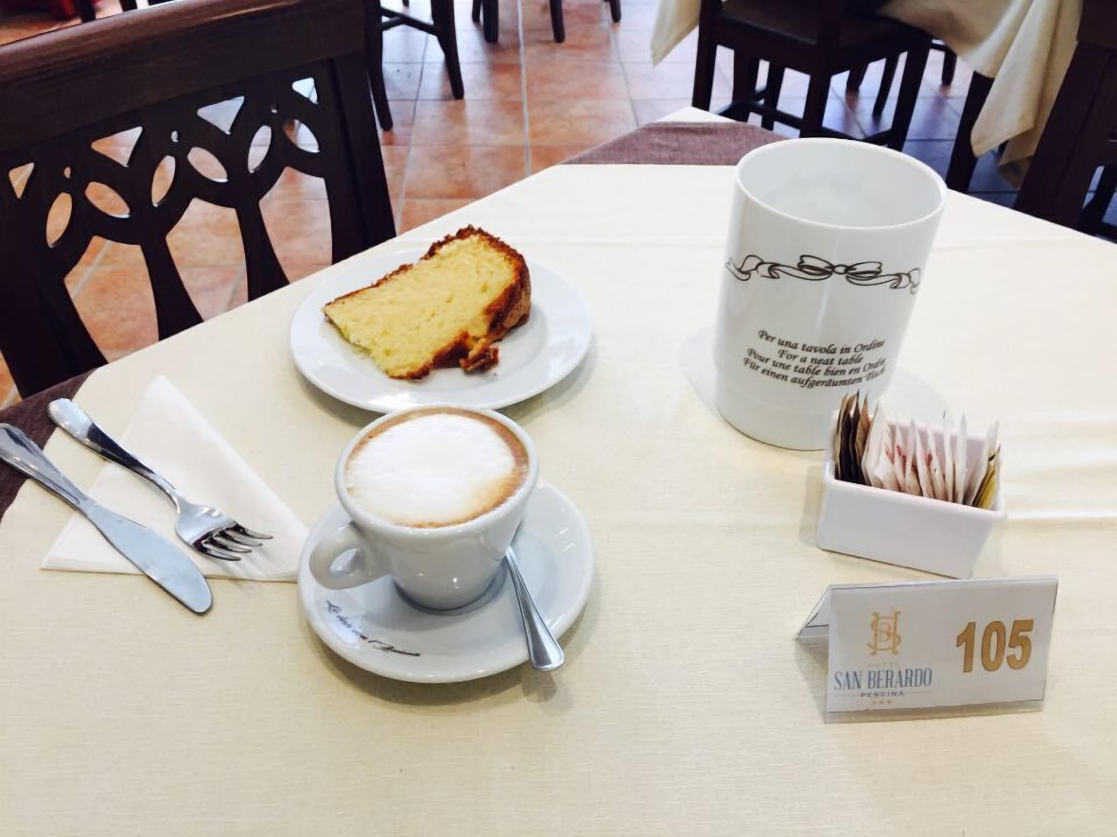 A table with a slice of cake, a cup of coffee, a mug, and a box labeled 'SAN BERARDO'.