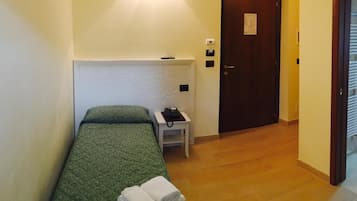 A single bed with a green bedspread, a bedside telephone, and a stack of white towels on the floor.