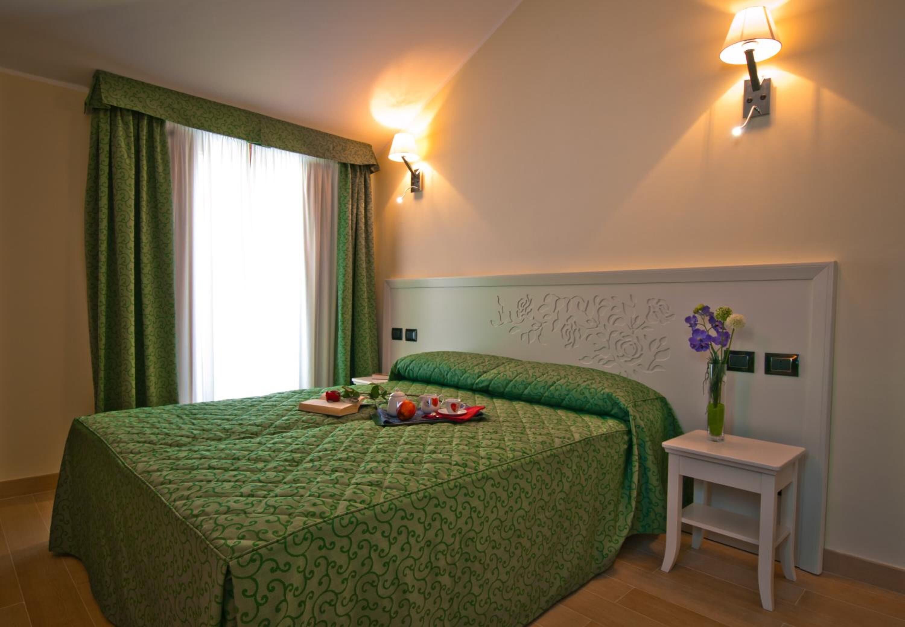 Habitación de hotel con cama, cortinas verdes, una mesita de noche con un florero y lámparas fijadas a la pared.
