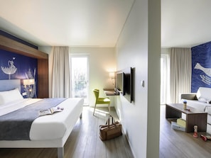 Quarto de hotel com uma cama, escrivaninha, televisão, cadeira e sofá.