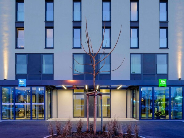 Un edificio moderno con un árbol frente a él, puertas de cristal y letreros que indican 'ibis budget' e 'ibis styles'.