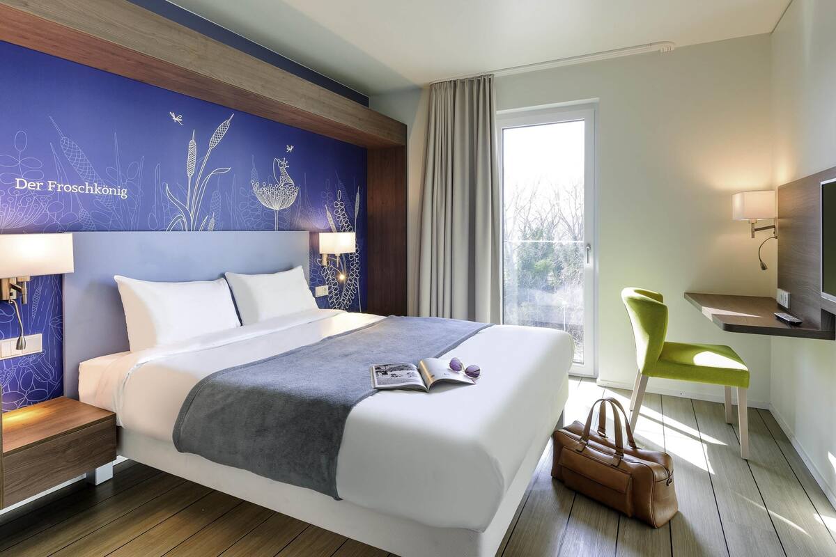A hotel room with a bed, bedside lamps, a desk with a chair, a television, and a window with curtains.