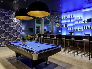 A pool table with surrounding chairs, a bar area with shelves of bottles, and a patterned wall. - ibis Styles Konstanz (Konstanz)