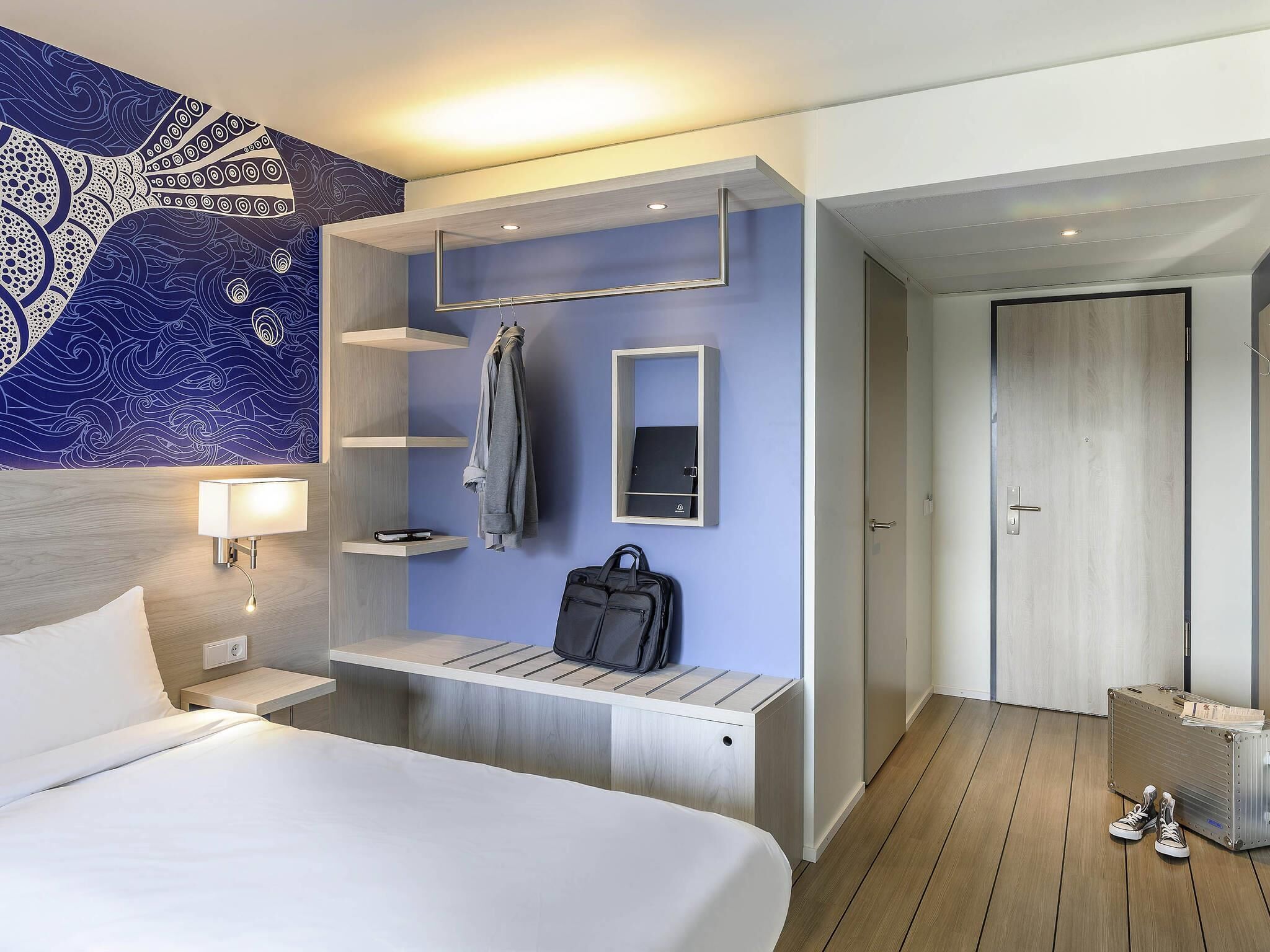 A modern hotel room with a bed, a wall-mounted desk, a coat rack, a suitcase, and a door.