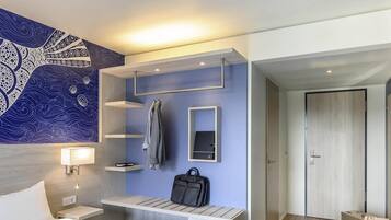 A modern hotel room with a bed, a wall-mounted desk, a coat rack, a suitcase, and a door.