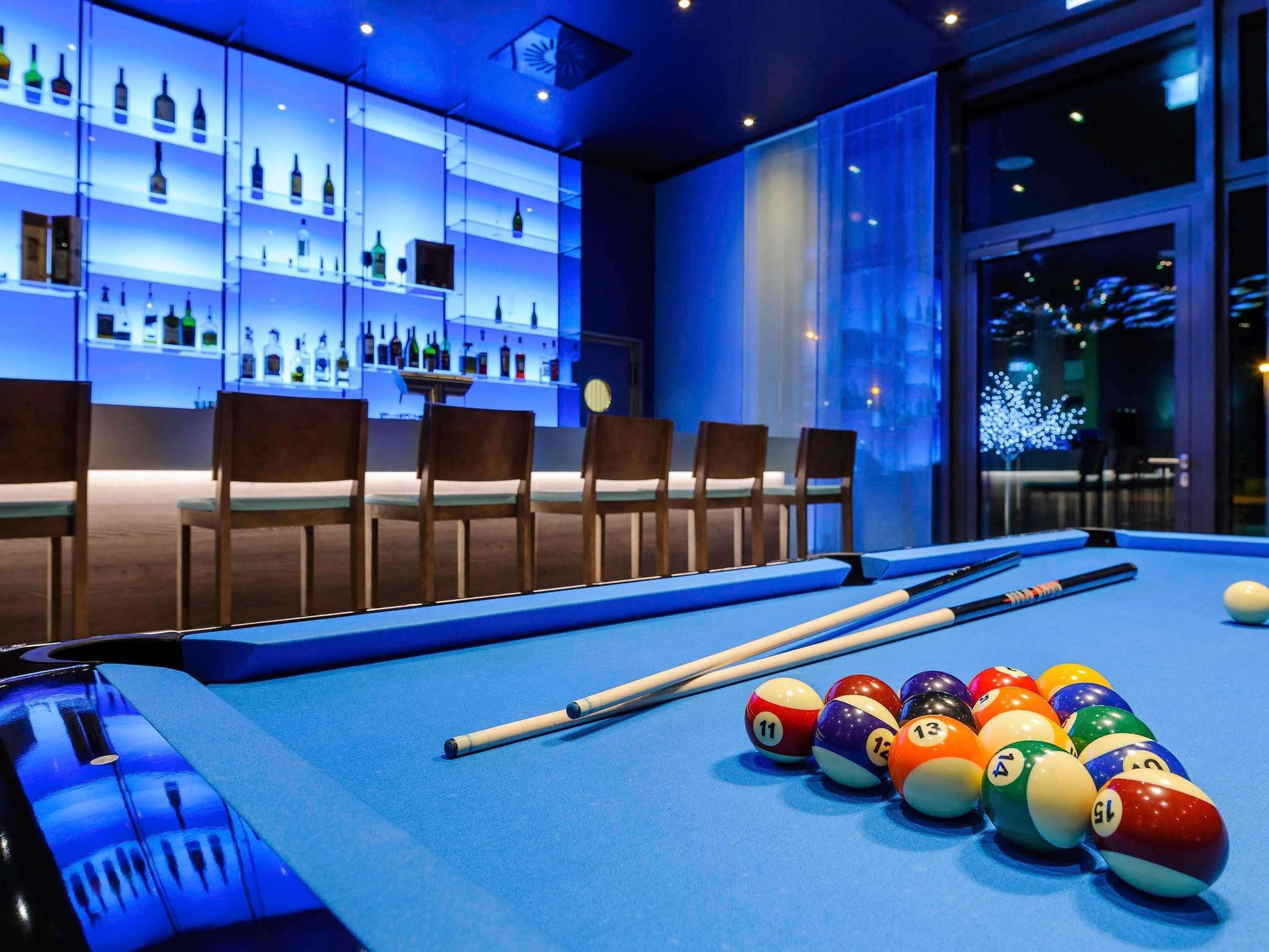 A billiards table with balls and cues, a bar area with shelves of bottles, and a seating arrangement with chairs.