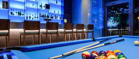 A billiards table with balls and cues, a bar area with shelves of bottles, and a seating arrangement with chairs.