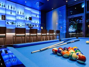 Une table de billard avec des billes et des queues, un bar avec des étagères remplies de bouteilles, et un espace détente avec des chaises.