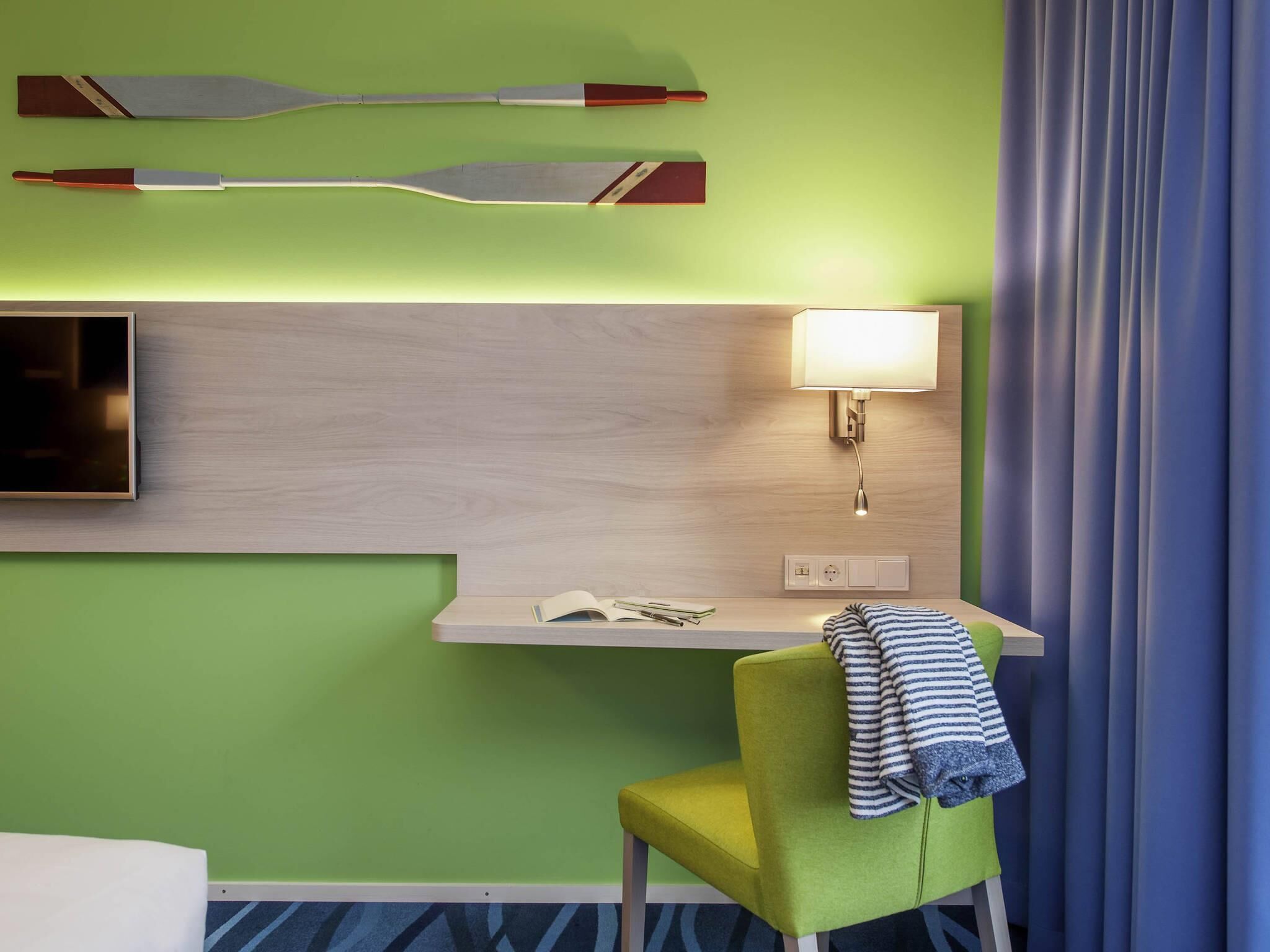 A hotel room with a green wall, a wooden desk with a lamp, a TV, and a striped towel on a yellow chair.