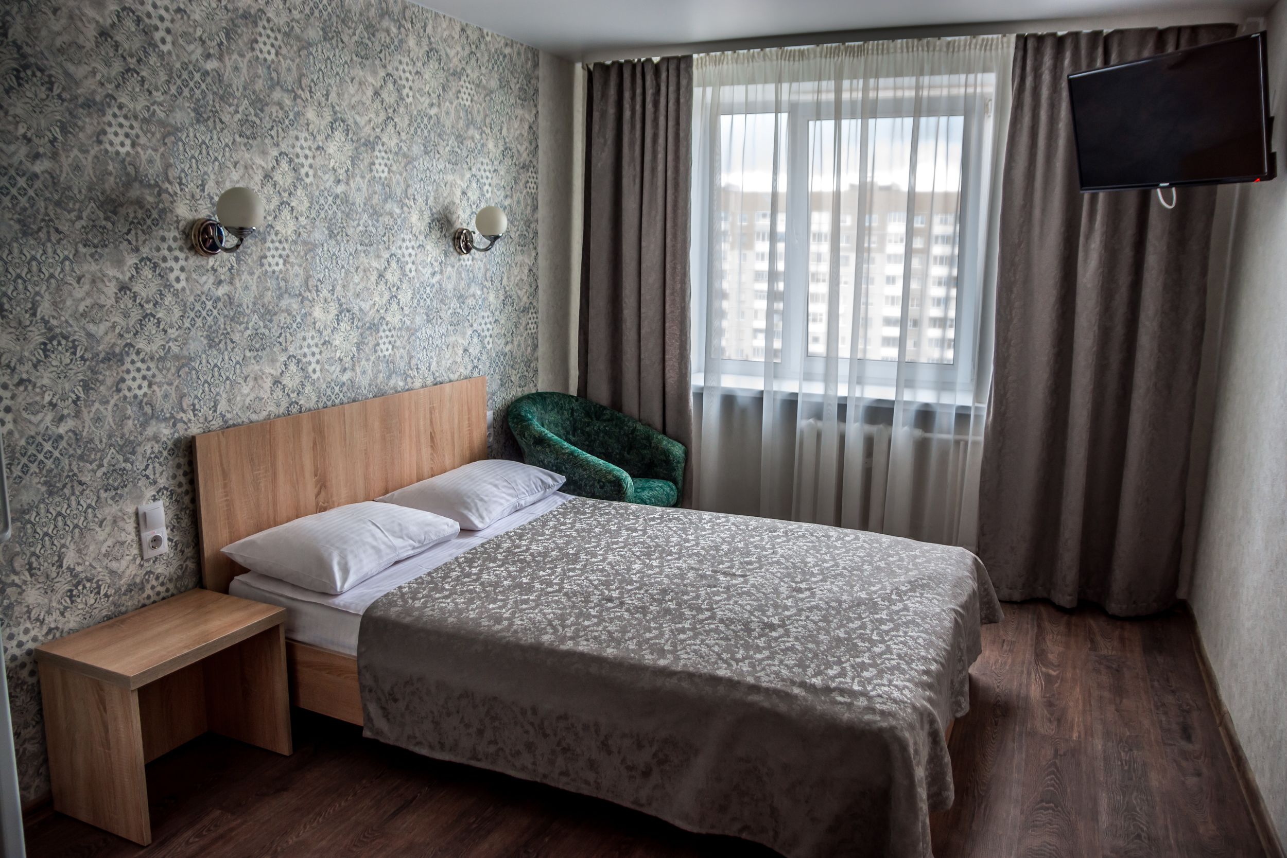 A hotel room with a bed, a nightstand, a green chair, a TV, and a window with curtains.