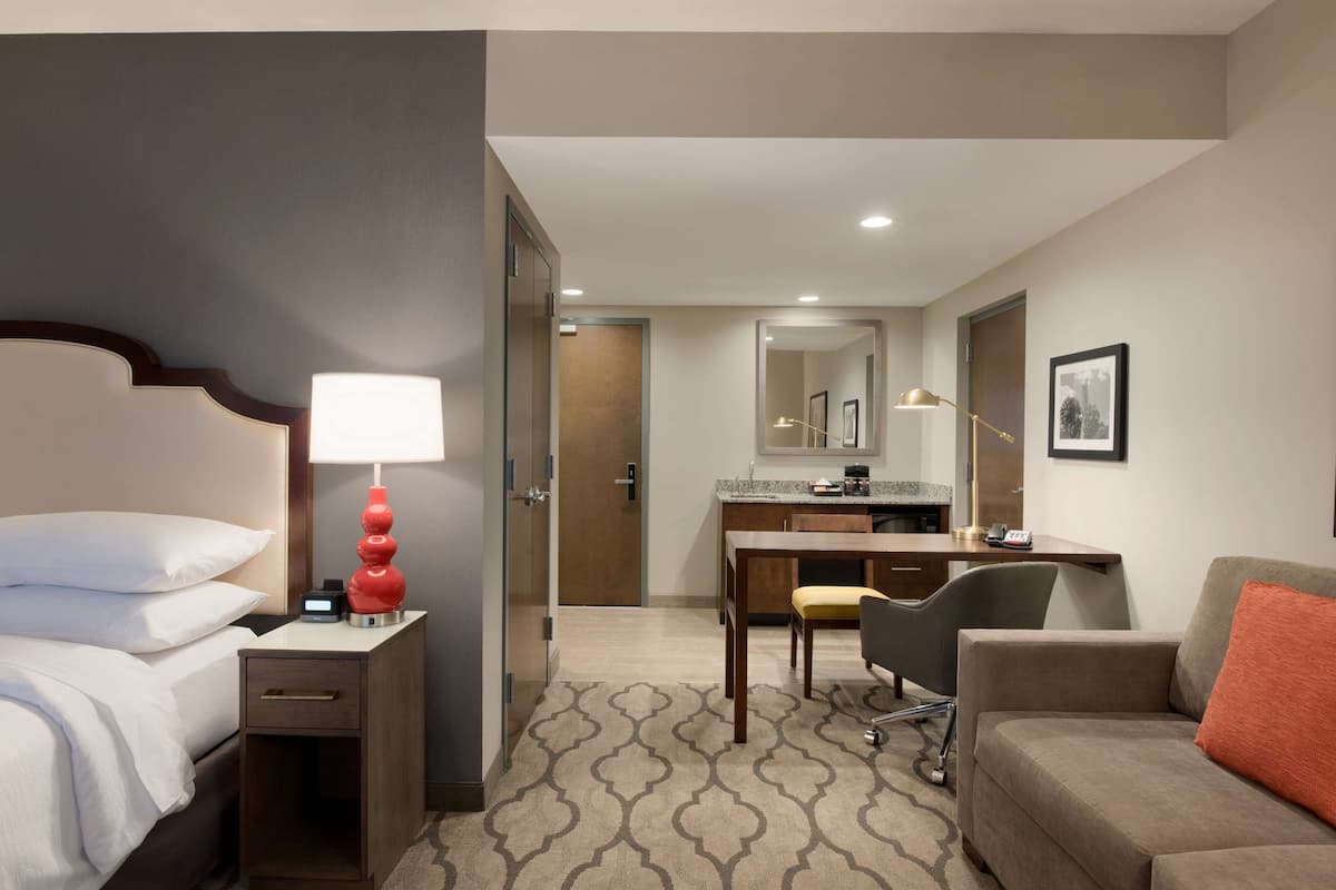 A hotel room with a bed, bedside table with a lamp, a desk with a chair, a sofa, and a kitchenette area.