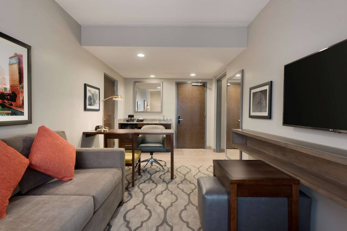 A hotel room with a grey sofa, a small wooden table, a TV mounted on the wall, a desk with a chair, and a large mirror.