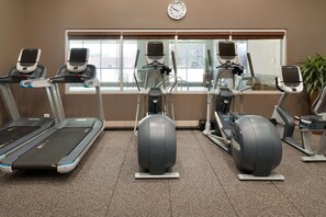 A gym with treadmills, elliptical machines, and a stationary bike, with a clock on the wall and a window with blinds.