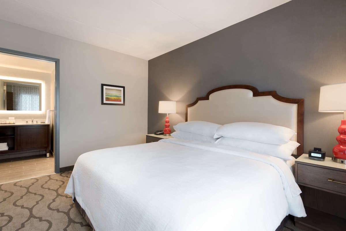 A hotel room with a large bed, a bedside table with a lamp, a nightstand, a mirror, and a bathroom area with a sink and toilet.