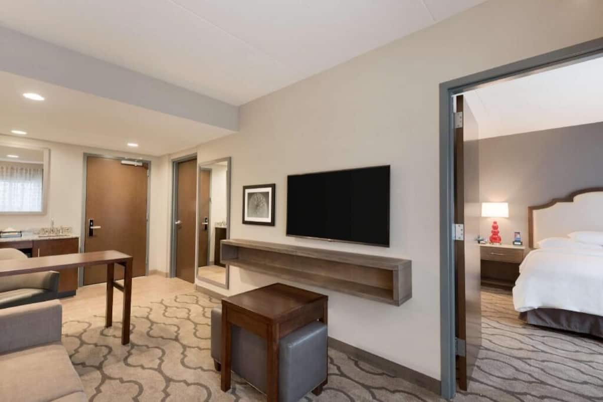 A hotel room with a bed, a TV mounted on the wall, a small table, and a seating area with a sofa and chair.