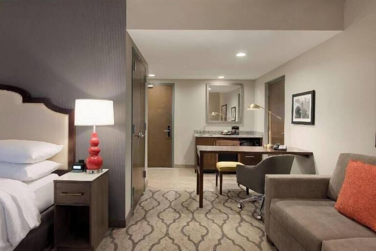 A hotel room with a bed, bedside table, lamp, desk, chair, sofa, and a small kitchenette.