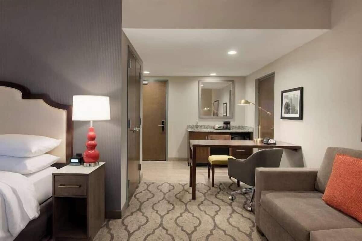 A hotel room with a bed, bedside table, lamp, desk, chair, and a sofa.